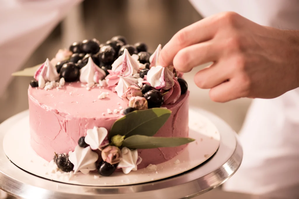 Confectioner decorating a pink wedding cake with meringues, berries, and floral accents.