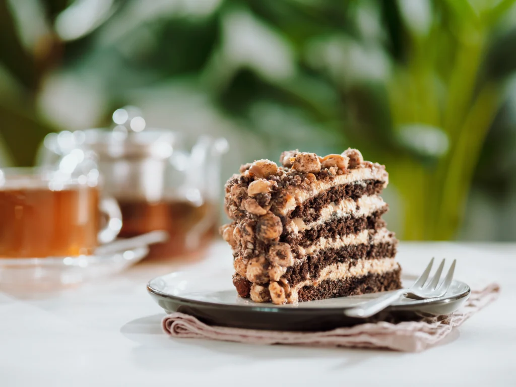 A rich, moist walnut cake with a golden-brown crust, topped with a layer of smooth chocolate ganache. The cake is adorned with finely chopped walnuts and whole walnut halves, adding both flavor and a delightful crunch. The interior is filled with ground walnuts, giving the cake a nutty aroma and a dense, buttery texture.