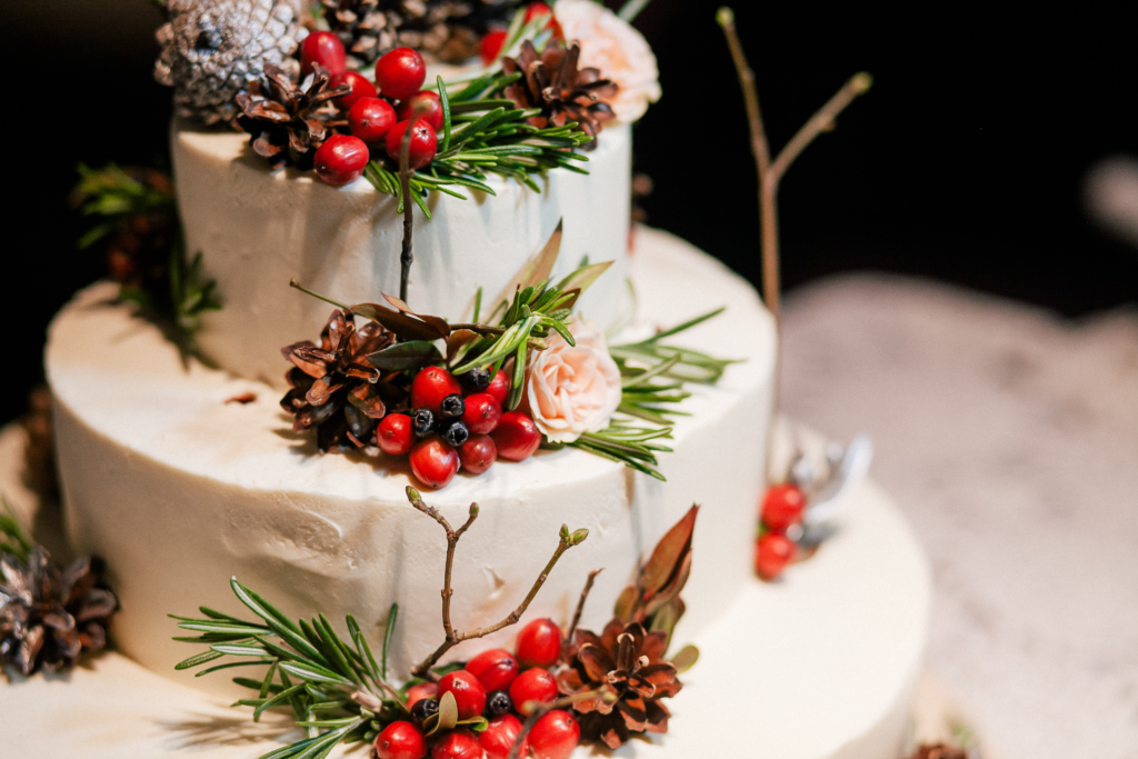 Three-tier winter wedding cake adorned with pine cones, red berries, rosemary sprigs, and pink roses.